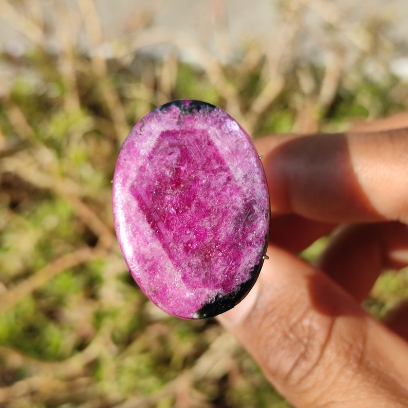 May include: An oval gemstone with a bright pink core and a dark edge. The stone's surface is polished, highlighting its natural patterns and textures. It's set in a thin metal frame, suitable for jewellery design. The gemstone is approximately 3 cm long.
