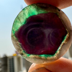 K&ouml;nnte beinhalten: Nahaufnahme eines runden, polierten Edelsteins. Der Stein hat ein tiefviolettes Zentrum mit wirbelnden Mustern, umgeben von leuchtendem Gr&uuml;n und einem hellen &auml;u&szlig;eren Rand. Der Edelstein wird zwischen Fingern gehalten.