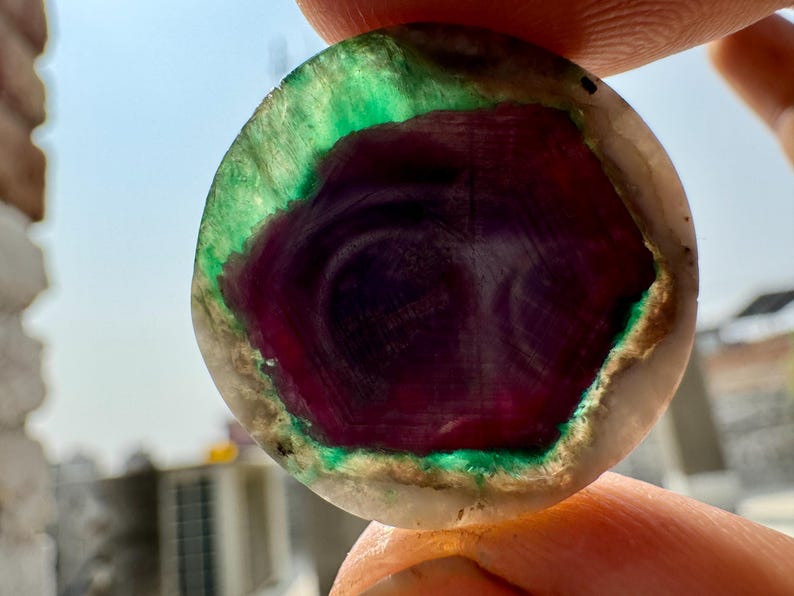 K&ouml;nnte beinhalten: Nahaufnahme eines runden, polierten Edelsteins. Der Stein hat ein tiefviolettes Zentrum mit wirbelnden Mustern, umgeben von leuchtendem Gr&uuml;n und einem hellen &auml;u&szlig;eren Rand. Der Edelstein wird zwischen Fingern gehalten.