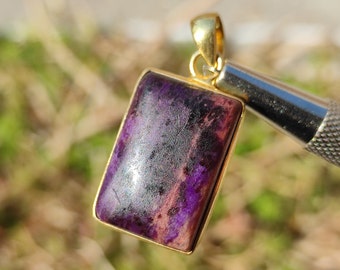 Pode incluir: Um pingente de ouro com uma pedra preciosa retangular de cor púrpura e preta. O pingente é engastado em uma moldura de ouro e tem um laço na parte superior para prender uma corrente.