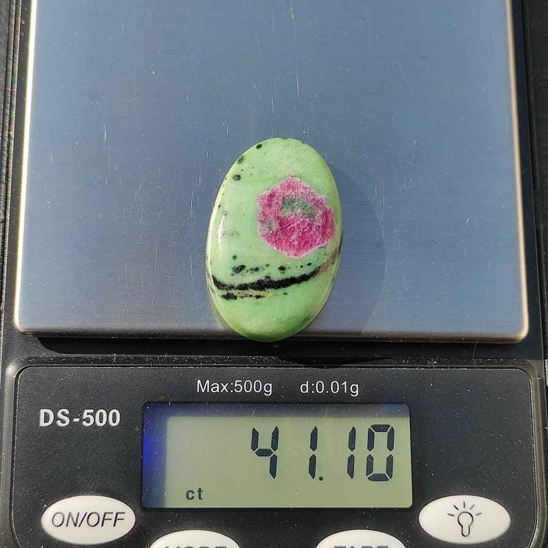 Peut inclure: Une pierre pr&eacute;cieuse ovale de couleur verte avec des marques noires et roses, pesant 4,11 carats, sur une balance num&eacute;rique.