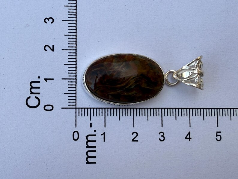 May include: Oval pendant with a brown and tan stone set in a silver-colored metal frame. The pendant measures approximately 1.5 inches in length. The stone has a marbled appearance. The bail is a decorative floral design.