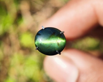 May include: A green gemstone with a chatoyant effect, also known as a cat's eye, is held in place by prongs. The stone is oval-shaped and has a light green sheen.