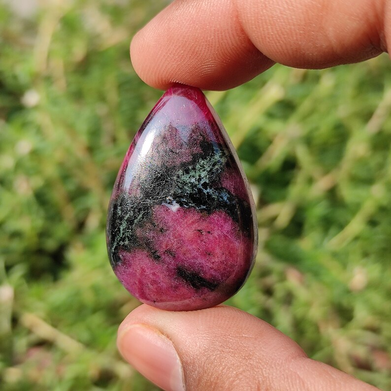Puede incluir: Un colgante de piedra preciosa cabuj&oacute;n en forma de l&aacute;grima con un patr&oacute;n de color rosa, negro y verde. El colgante est&aacute; hecho de zoisita rub&iacute;.