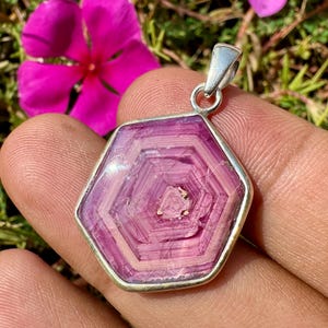 May include: A hexagonal pendant featuring a pink tourmaline slice with concentric geometric patterns, encased in a polished silver metal frame with a top bail, showcasing natural crystalline layers and a vibrant magenta hue.