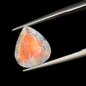 May include: A teardrop-shaped gemstone with a pearlescent finish. The gemstone is a pale pink color with iridescent flashes of color. It is held in tweezers against a black background.