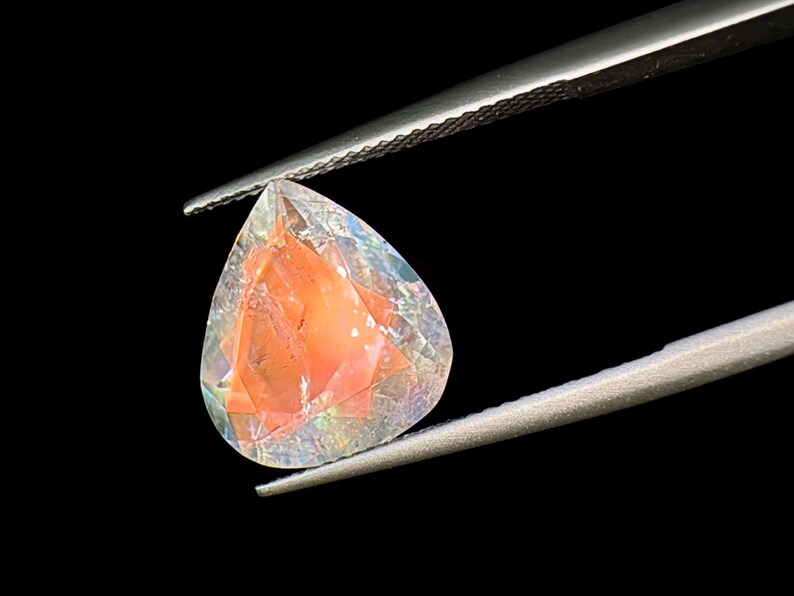 May include: A teardrop-shaped gemstone with a pearlescent finish. The gemstone is a pale pink color with iridescent flashes of color. It is held in tweezers against a black background.
