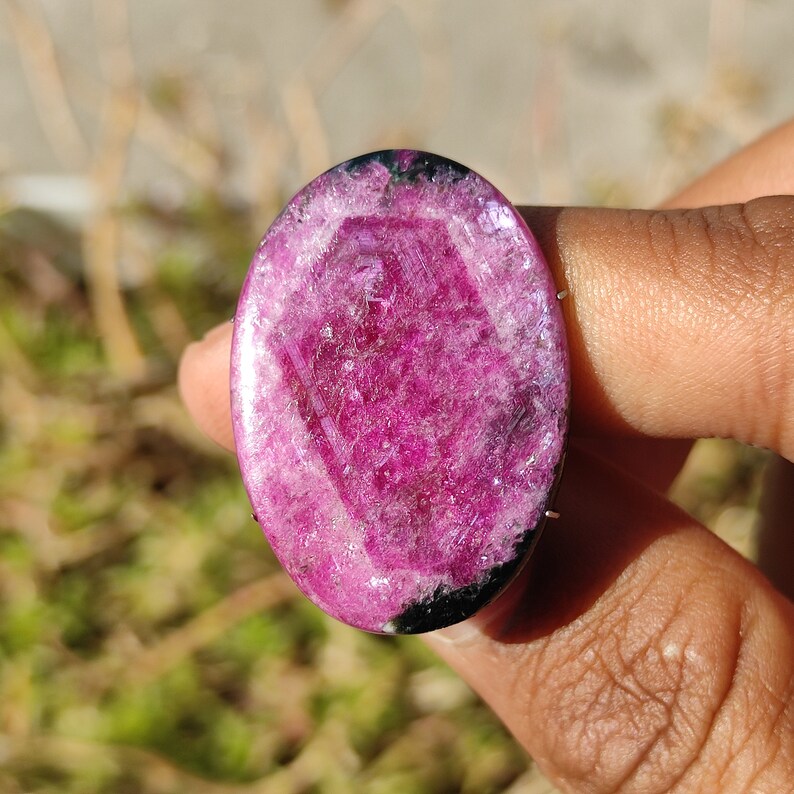 May include: An oval-shaped, polished gemstone in a vibrant magenta colour, featuring a central geometric pattern. The stone has a translucent quality, with areas of black and white. The gemstone is held in a hand, with a blurred green background.
