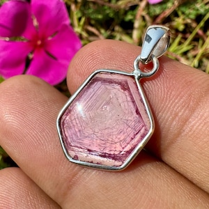 May include: A hexagonal pendant featuring a translucent pink gemstone with visible internal geometric patterns, encased in a polished silver-toned metal setting with a small circular bail at the top for attachment to a chain.