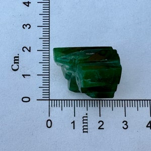May include: A dark green, faceted, crystal-like rock with a rough, uneven surface. The rock is about 2 cm wide and 1 cm tall.