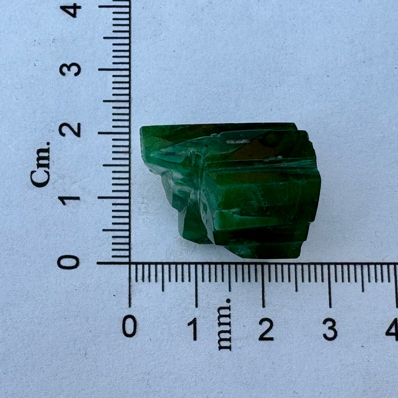 May include: A dark green, faceted, crystal-like rock with a rough, uneven surface. The rock is about 2 cm wide and 1 cm tall.
