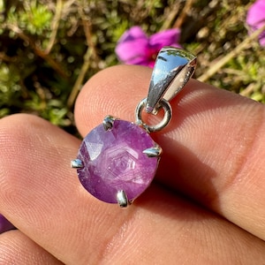 May include: A round, faceted, purple gemstone pendant with a silver bail. The gemstone is held in place by four silver prongs. The pendant is displayed against a background of green foliage and pink flowers.