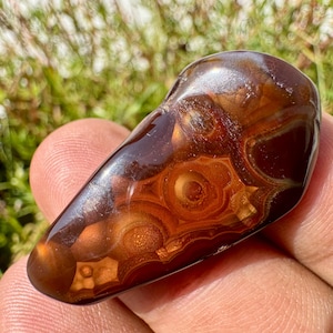 May include: A polished, teardrop-shaped fire agate gemstone featuring deep reddish-brown tones and translucent, glowing orange orb-like inclusions. The surface is smooth and reflective, showcasing intricate, layered banding patterns throughout the stone.