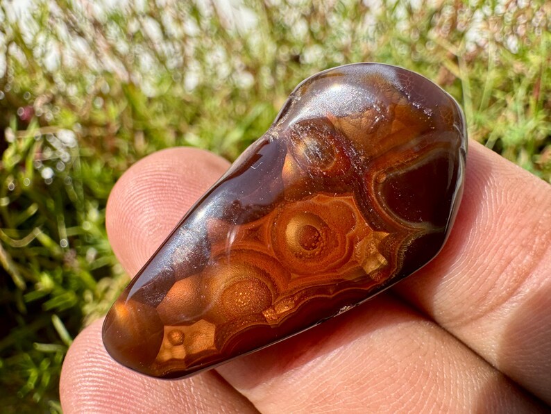 May include: A polished, teardrop-shaped fire agate gemstone featuring deep reddish-brown tones and translucent, glowing orange orb-like inclusions. The surface is smooth and reflective, showcasing intricate, layered banding patterns throughout the stone.