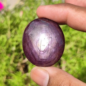 May include: A deep purple, round, cabochon cut gemstone with a star-like pattern. The gemstone is held in a hand.