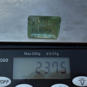 Op de afbeelding: Een rechthoekige, doorschijnende, lichtgroene edelsteen ligt op een digitale weegschaal. De weegschaal geeft 23,75 ct aan. De weegschaal, model DS-500, heeft een maximale capaciteit van 500g en een precisie van 0,01g.
