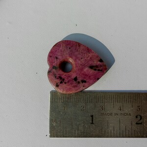 Op de afbeelding: Een roze hartvormige steen met zwarte vlekken en een gat in het midden. De steen is ongeveer 5 cm lang en ligt op een liniaal.