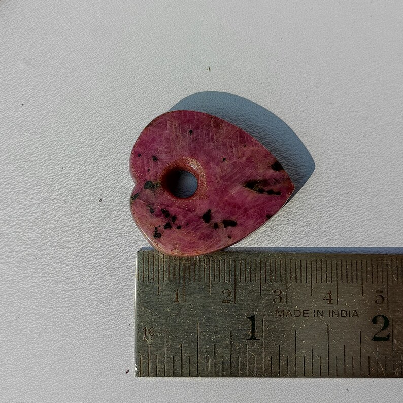 Op de afbeelding: Een roze hartvormige steen met zwarte vlekken en een gat in het midden. De steen is ongeveer 5 cm lang en ligt op een liniaal.