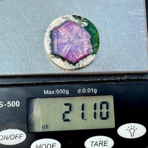 K&ouml;nnte beinhalten: Ein runder Edelstein mit einem lila und wei&szlig;en Muster, umgeben von Gr&uuml;n und Wei&szlig;, liegt auf einer Digitalwaage. Die Waage zeigt ein Gewicht von 21,10 ct an. Die Waage ist schwarz mit wei&szlig;em Text und Kn&ouml;pfen.