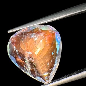 May include: A heart-shaped gemstone with a clear, faceted body and a reddish-brown interior. The gemstone is held by a pair of tweezers.