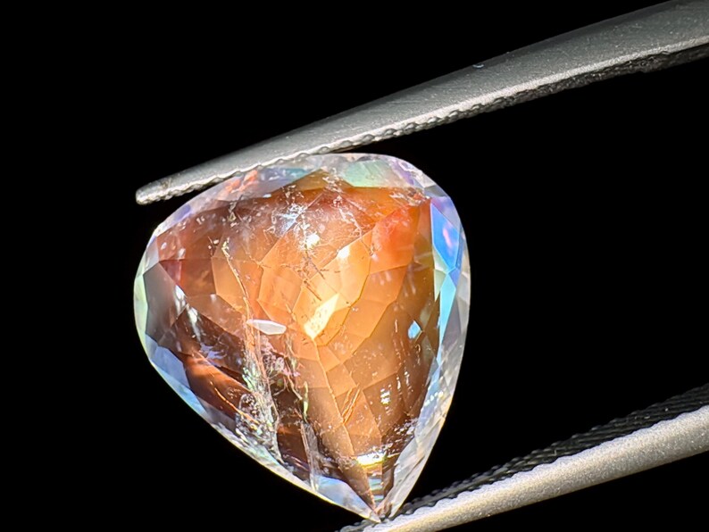 May include: A heart-shaped gemstone with a clear, faceted body and a reddish-brown interior. The gemstone is held by a pair of tweezers.