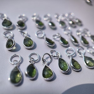 Op de afbeelding: Een verzameling zilveren hangers in de vorm van een traan met groene edelsteenaccenten. De hangers zijn gerangschikt op een wit oppervlak.