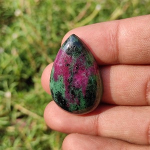May include: A teardrop-shaped green, pink, and black gemstone cabochon. The gemstone is a natural stone with a smooth, polished surface.