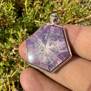 May include: A hexagonal pendant featuring a purple star ruby gemstone set in sterling silver. The ruby displays a star-like pattern and has a polished, shiny finish.