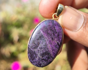 Pode incluir: Um pingente oval roxo e dourado com uma argola dourada. O pingente é feito de uma pedra natural com um padrão de redemoinho.