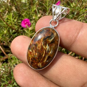 May include: Oval pendant with a polished tiger's eye stone, featuring a silver-colored bail. The stone displays a mix of brown, gold, and yellow hues. The pendant is held in a hand, with a green and pink floral background.