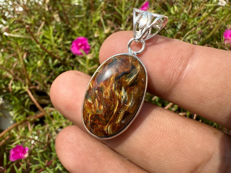 May include: Oval pendant with a polished tiger's eye stone, featuring a silver-colored bail. The stone displays a mix of brown, gold, and yellow hues. The pendant is held in a hand, with a green and pink floral background.