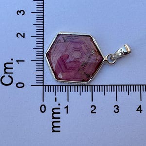 K&ouml;nnte beinhalten: Ein sechseckiger, rosafarbener Edelsteinanh&auml;nger in einer silbernen Fassung, platziert neben einem Lineal, das zeigt, dass der Anh&auml;nger etwa 2 cm breit und 1,5 cm hoch ist. Der Stein weist ein sichtbares konzentrisches Sechseckmuster auf.