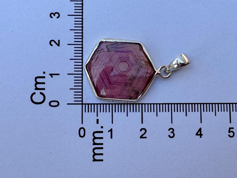 K&ouml;nnte beinhalten: Ein sechseckiger, rosafarbener Edelsteinanh&auml;nger in einer silbernen Fassung, platziert neben einem Lineal, das zeigt, dass der Anh&auml;nger etwa 2 cm breit und 1,5 cm hoch ist. Der Stein weist ein sichtbares konzentrisches Sechseckmuster auf.