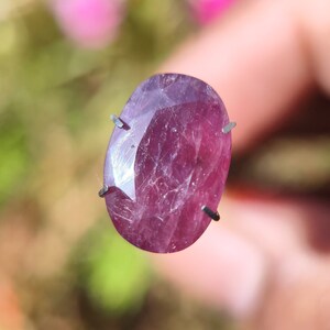 May include: A deep pink oval-shaped gemstone with a faceted cut. The gemstone is held in place by prongs on a metal stand.