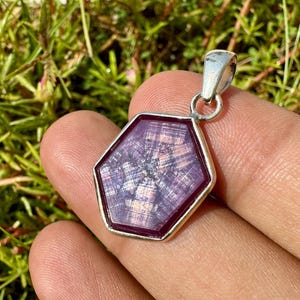 Könnte beinhalten: Ein sechseckiger Anhänger mit einem violetten Sternsaphir-Edelstein in einer silberfarbenen Metallfassung. Der Edelstein zeigt ein ausgeprägtes sechsstrahliges Sternmuster auf seiner Oberfläche, mit polierter Oberfläche und einer silbernen Öse.