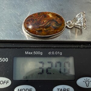 May include: An oval pendant with a silver setting and a polished gemstone. The gemstone has a swirling pattern of brown, orange, and black. The pendant is on a digital scale, which reads 32.70 ct.