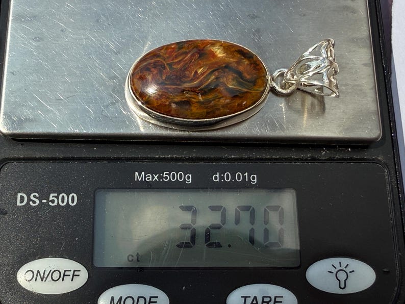 May include: An oval pendant with a silver setting and a polished gemstone. The gemstone has a swirling pattern of brown, orange, and black. The pendant is on a digital scale, which reads 32.70 ct.