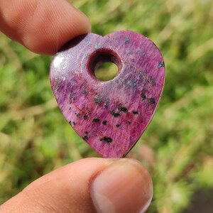 May include: A heart-shaped pendant made of pink ruby with black inclusions. The pendant has a hole in the center for a cord or chain.