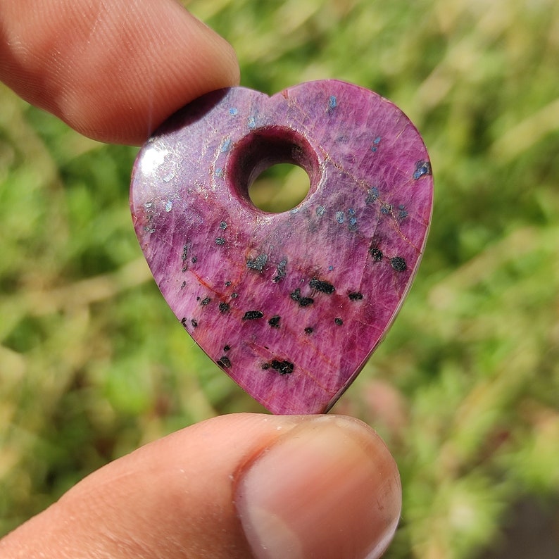 May include: A heart-shaped pendant made of pink ruby with black inclusions. The pendant has a hole in the center for a cord or chain.