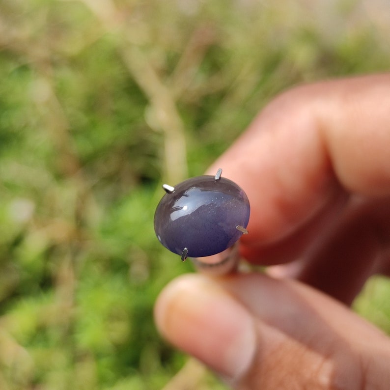 May include: A close-up of a dark blue oval-shaped gemstone with a star-like pattern, held in a prong setting.