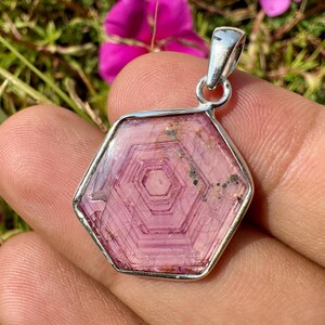 K&ouml;nnte beinhalten: Ein sechseckiger Anh&auml;nger mit einem durchscheinenden rosa Edelstein mit konzentrischen geometrischen Mustern, gefasst in einen polierten Silberrahmen mit einer silbernen &Ouml;se an der Oberseite und nat&uuml;rlichen Einschl&uuml;ssen.
