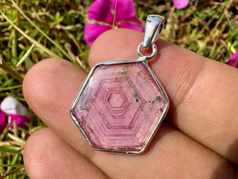 K&ouml;nnte beinhalten: Ein sechseckiger Anh&auml;nger mit einem durchscheinenden rosa Edelstein mit konzentrischen geometrischen Mustern, gefasst in einen polierten Silberrahmen mit einer silbernen &Ouml;se an der Oberseite und nat&uuml;rlichen Einschl&uuml;ssen.