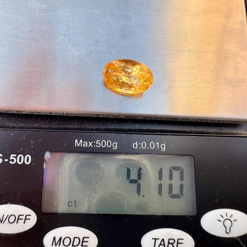 May include: A close-up of a yellow-orange gemstone on a digital scale. The scale reads 4.10 ct.