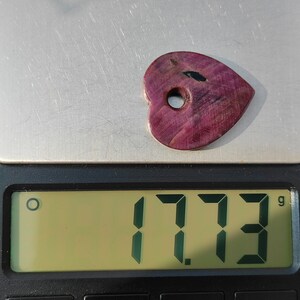 以下が含まれることがあります： 黒いインクルージョンが入ったハート型の紫色の宝石で、重さは1.773グラムです。宝石には穴が開いており、ペンダントやチャームであることを示唆しています。