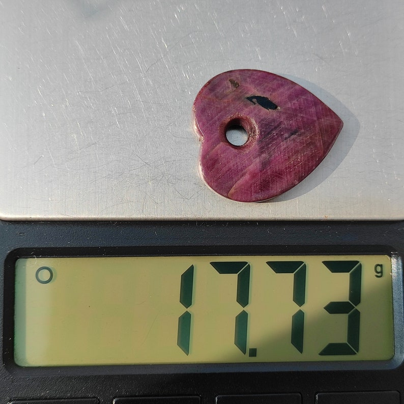 以下が含まれることがあります： 黒いインクルージョンが入ったハート型の紫色の宝石で、重さは1.773グラムです。宝石には穴が開いており、ペンダントやチャームであることを示唆しています。