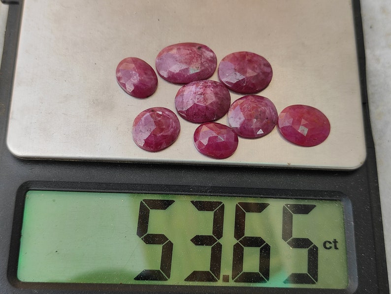 Pu&ograve; includere: Primo piano di una bilancia digitale che mostra 5,36 carati. La bilancia mostra una collezione di 9 pietre preziose di rubino sfaccettate, di diverse dimensioni e forme. I rubini sono di colore rosa intenso.