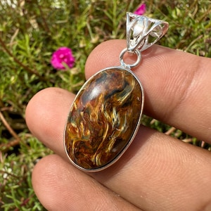 May include: Oval pendant with a polished tiger's eye stone, showcasing a blend of brown and gold hues. The stone is set in a silver-colored metal frame with a decorative bail. The pendant is held in a hand against a backdrop of green foliage and pink flowers.