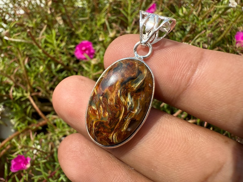 May include: Oval pendant with a polished tiger's eye stone, showcasing a blend of brown and gold hues. The stone is set in a silver-colored metal frame with a decorative bail. The pendant is held in a hand against a backdrop of green foliage and pink flowers.