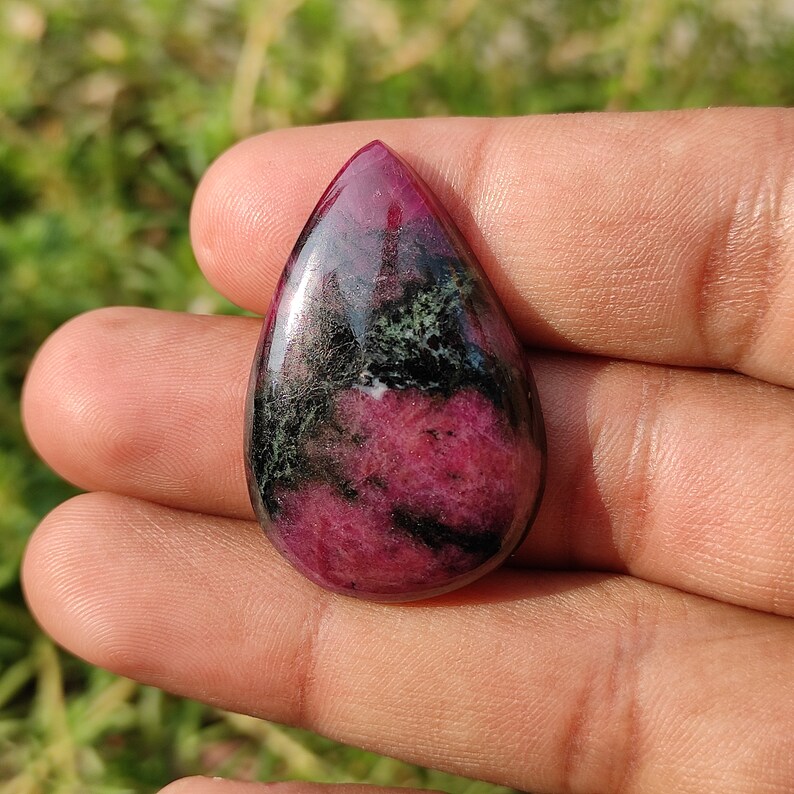 Puede incluir: Un colgante de piedra preciosa cabuj&oacute;n en forma de l&aacute;grima con un patr&oacute;n de color rosa y verde. La piedra tiene una superficie lisa y pulida y est&aacute; engastada en un engaste de tono plateado.