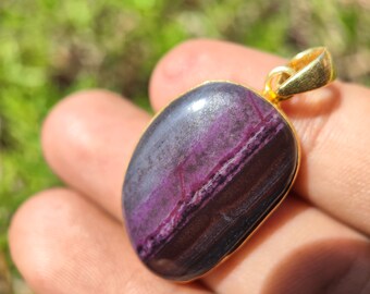 Pode incluir: Um pingente de ouro com uma pedra listrada de roxo e preto. A pedra tem formato oval e um acabamento liso e polido.